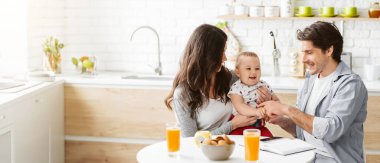 Bu resim, mutlu bir aileyi kahvaltıda birlikte mutfaklarında gösteriyor. Anne bebeğini kucağında tutuyor, o da gülümsüyor ve babasına bakıyor, boşluğu kopyalıyor.