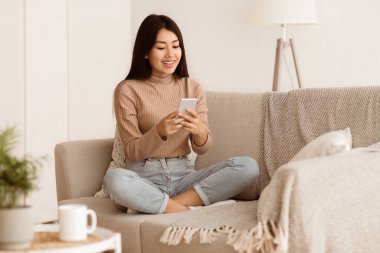 Genç bir kadın oturma odasındaki bir kanepede oturuyor ve telefon kullanıyor. Gülümsüyor ve telefona bakıyor..