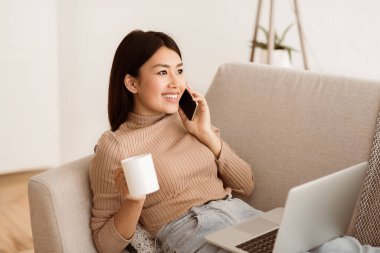 Kanepede oturan bir kadın telefonla konuşurken gülümsüyor. Diğer elinde bir kupa kahve tutuyor ve önünde bir dizüstü bilgisayar açık..