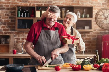 Neşeli yaşlı bir çift mutfaklarında doğum gününü kutluyor. Kadın, erkeğin gözünün arkasında paketlenmiş bir hediyeyle yemek hazırlıyor. Görüntü rahat ve sevgi dolu bir atmosferi gösteriyor..