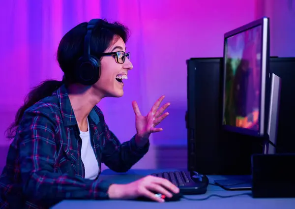 stock image The image shows a woman wearing glasses and headphones, smiling and excitedly playing video games on her computer late at night. She has her hands on the keyboard and mouse