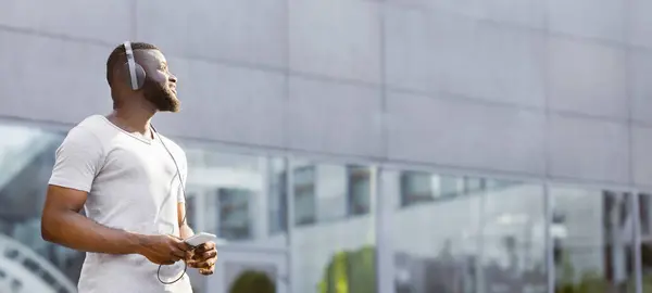 Stock image Music lover. Young serene african american guy walking outdoor in headphones, panorama, free space