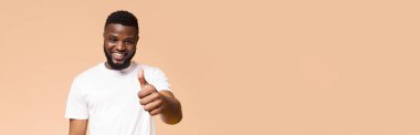 Kısa, siyah saçlı ve sakallı gülümseyen siyah bir adam beyaz bir tişört giyiyor. O, sağ eliyle baş parmak işareti yapıyor. Arkaplan şeftali renginde, boşluğu kopyala