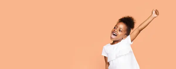 stock image A little black girl with curly hair and a bright smile raises her arm in celebration against a peach-colored background. She is wearing a white t-shirt and appears to be full of joy and excitement