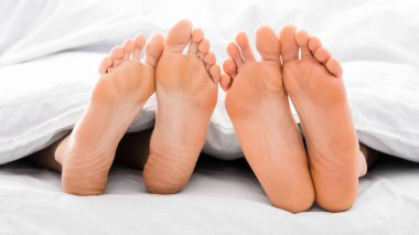 Beyaz çarşafların altından sarkan iki çift çıplak ayakla yakın çekim. Ayaklar yan yana, parmaklar kamerayı işaret ediyor.