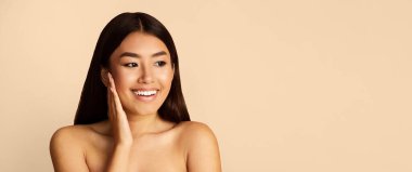 Uzun siyah saçlı genç bir Asyalı kadın eliyle yanağına dokunurken geniş bir gülümseme sergiliyor. Yan tarafa bakıyor ve arka plan şeftali rengi, fotokopi alanı, panorama.