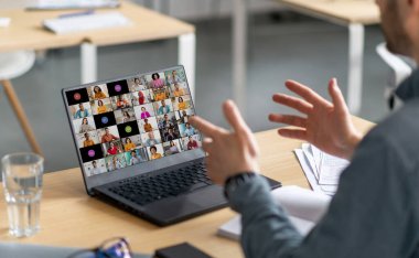 Bir adam dizüstü bilgisayarın önünde bir masada oturuyor. Elleriyle el kol hareketi yapıyor ve ekranda birden fazla kişiyle görüntülenen görüntülere bakıyor..