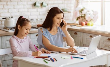 Bir kadın kızıyla mutfak masasında oturuyor. Kadın dizüstü bilgisayarda çalışıyor ve telefonla konuşuyor. Kızı bir kağıt parçasını boyuyor..