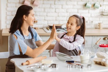 Bir anne ve küçük kızı mutfakta yemek pişirirken gülümsüyor ve beşlik çakıyorlar. Mutfak temiz ve modern ve masada bir sürü pişirme malzemesi var.