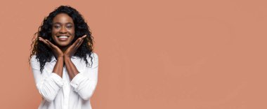 Kıvırcık saçlı, gülümseyen siyah bir kadın şeftali rengi bir arka plana poz veriyor. Ellerini yüzüne dolamış, mutlu ve kaygısız görünüyor. Beyaz düğmeli bir gömlek giyiyor..