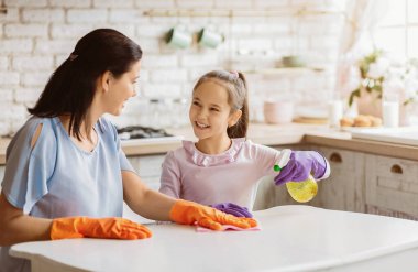 Bir anne ve kızı birlikte bir mutfak masasını temizliyorlar. Anne turuncu eldiven giyiyor ve masayı bir bezle siliyor. Kız mor eldivenler giyiyor ve masayı ilaçlıyor.