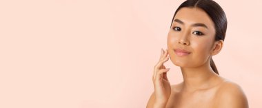 Uzun siyah saçlı genç bir Asyalı kadın yüzüne dokunurken nazikçe gülümsüyor. Açık pembe bir arka planın karşısında duruyor. Cildi pürüzsüz ve sağlıklı görünüyor, kopyalama alanı