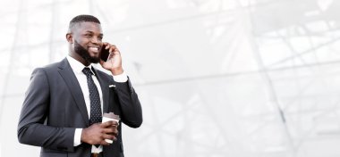 Gülümseyen Afro İş Adamı cep telefonuyla konuşuyor elinde kahve fincanıyla modern binanın yanında yürüyor. Boşluğu Kopyala