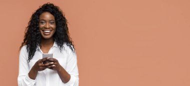 Akıllı telefonu elinde tutarken kıvırcık saçlı genç bir zenci kadın gülümsüyor. Beyaz düğmeli bir gömlek giyiyor ve şeftali arkaplanının önünde duruyor..