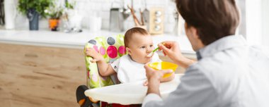 Bir baba, bebeğini bir mutfakta sandalyede besler. Bebek beyaz bir önlük giyiyor ve renkli bir yüksek sandalyede oturuyor. Babası bebeği beslemek için kaşık kullanıyor.