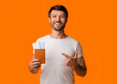 Beyaz tişörtlü gülümseyen bir adam sol elinde bir pasaport ve uçak bileti tutuyor ve sağ elinde parlak turuncu bir arka planla sağ elini gösteriyor..