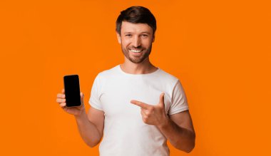 Beyaz tişörtlü bir adam elinde beyaz ekranlı siyah bir akıllı telefon tutuyor ve ona gülümsüyor, turuncu bir arka planda duruyor..