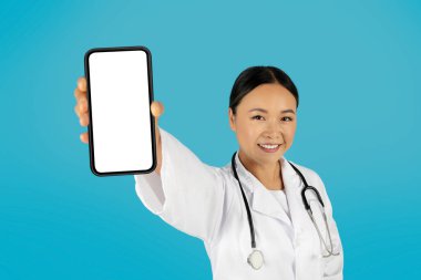 Beyaz önlüklü Asyalı kadın doktor ve steteskop kameraya gülümserken sağ elinde boş bir ekranla akıllı telefonu tutuyor. Arka plan parlak bir mavi..