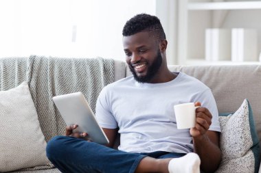 Siyah adam bir elinde dijital tablet, diğer elinde bir kupa kahve tutarak kanepede rahatça oturur. Tabletteki bir şeye bulaşmış gibi gülümsüyor..