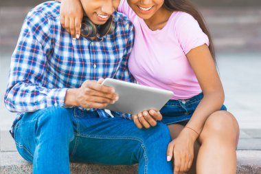 Genç bir Afro-Amerikan çift açık hava basamaklarında oturup birlikte bir tablet cihazına bakıyorlar. Kız pembe bir gömlek ve kot etek giyiyor. Kolu da erkeklerin omuzlarında.