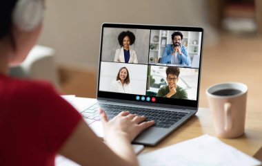 Önünde dizüstü bilgisayarı olan bir kadın, sırtı kameraya dönük bir şekilde masada oturuyor. Laptop ekranında görünen birçok katılımcıyla bir video konferans görüşmesine katılıyor.