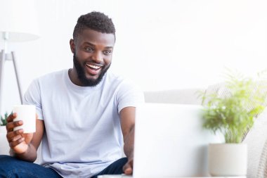 Afro-Amerikalı adam masasında oturuyor, dizüstü bilgisayarına bakarken gülümsüyor. Elinde beyaz bir fincan kahve tutuyor ve evden çalışıyormuş gibi görünüyor, kopyalama alanı.