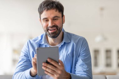 Bir adam tablet kullanırken gülümsüyor. Açık mavi düğmeli bir gömlek giyiyor ve içeride oturuyor. Cihazı kullanarak eğleniyor gibi görünüyor..
