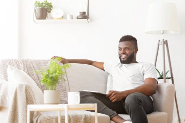 Beyaz tişörtlü ve siyah pantolonlu siyah bir adam, parlak ışıklandırılmış oturma odasındaki krem renkli bir kanepede rahatça oturur. Oda bir saksı çiçeği ve bir lambayla süslenmiş.