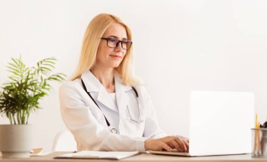 Beyaz önlüklü sarışın bir doktor ofisinde dizüstü bilgisayar kullanarak bir masada oturuyor..