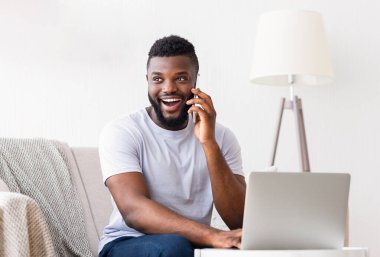 Sakallı genç siyah bir adam telefonuyla konuşurken gülümsüyor ve bir dizüstü bilgisayara bakıyor. Modern bir oturma odasında, arka planda beyaz bir lamba olan bir kanepede oturuyor..