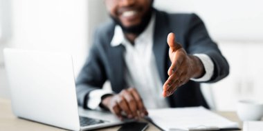 Gülümseyen bir iş adamı masasında oturur ve kamera önünde olmayan birine el sıkışma teklif eder..