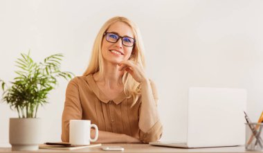 Gözlüklü sarışın bir kadın bir masada oturuyor, gülümsüyor, dizüstü bilgisayarı, bir fincan kahvesi ve arka planda bir bitkisi var..