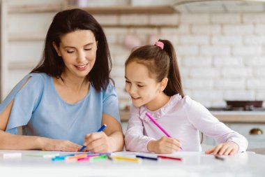 Bir anne ve kız mutfakta bir masada oturuyor, ikisi de dikkatle boyama projelerine odaklanıyor. Renkli kalemler ve pastel kalemler kullanıyorlar. Eğleniyor ve bağ kuruyorlar.