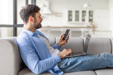 Modern bir evdeki bir kanepede oturan bir adam, rahat bir şekilde akıllı telefon ve dizüstü bilgisayar kullanıyor. Gülümsüyor, muhtemelen dijital bir işle meşgul ya da eğleniyor..