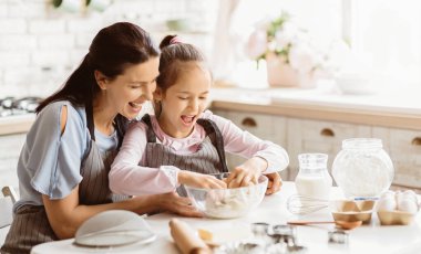 Bir anne ve küçük kızı mutfakta birlikte yemek pişiriyorlar. Malzemeleri karıştırmak için birlikte çalışırken ikisi de gülümsüyor ve eğleniyorlar. Mutfak aydınlık.