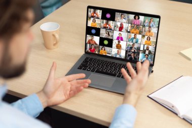 Bir adam bir dizüstü bilgisayarın önünde oturmuş, aktif olarak iş arkadaşlarıyla bir video konferansına katılıyor. Elleri havada, işaret ediyor ve online katılımcılarla iletişim kuruyor..