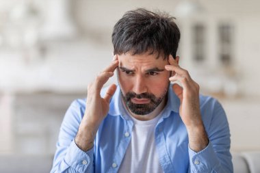 Görüntü, sakallı ve kısa siyah saçlı, açık mavi düğmeli gömlek giyen, elleri şakaklarının üzerinde bir evde oturan bir adamı gösteriyor. Endişeli bir ifadesi var, yakın çekim.