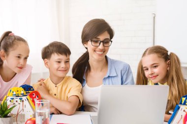 Bir öğretmen ve üç genç öğrenci bir sınıf ortamında dizüstü bilgisayarın etrafında toplanırlar. Robot bilimi öğreniyor gibi görünüyorlar. Öğretmen gülümsüyor ve ekranı işaret ediyor.