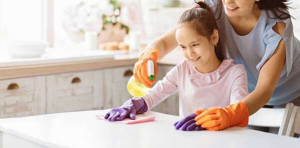 A young girl wearing pink shirt and purple gloves is cleaning a table in the kitchen while her mother, wearing a blue shirt and orange gloves, assists her, panorama with copy space