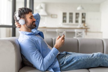 Bir adam kanepede oturuyor, kulaklık takıyor ve akıllı telefonunu kullanıyor. Rahatlamış ve zamanının tadını çıkarıyor gibi görünüyor. Adam açık mavi gömlek ve kot pantolon giyiyor.