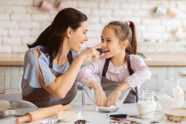 Bir anne ve kızı mutfakta birlikte yemek pişiriyorlar. Anne mavi bir gömlek ve çizgili bir önlük giyer, kız ise pembe bir gömlek ve çizgili bir önlük giyer.