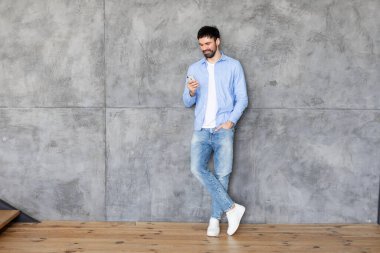 Mavi düğmeli gömlekli ve kot pantolonlu bir adam bir bacağı beton bir duvara çarpmış, akıllı telefonuna hafifçe gülümseyerek bakıyor. Adamın sakalı ve kısa koyu renk saçı var.