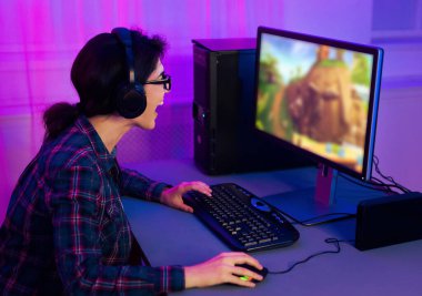 Bu resim ekose gömlek ve kulaklık takan genç bir kadını bilgisayarda video oyunu oynarken gösteriyor. O, bir klavye ve fareyle bir masada oturuyor..