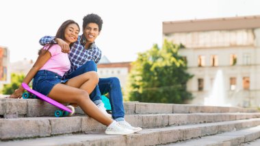 Afro-Amerikalı genç çift şehir parkında merdivenlerde oturuyor. Kız bacaklarını açık bir şekilde pembe bir kaykayın üzerinde oturuyor. Adamın kolu kızın omzunda ve ikisi de gülümsüyor.