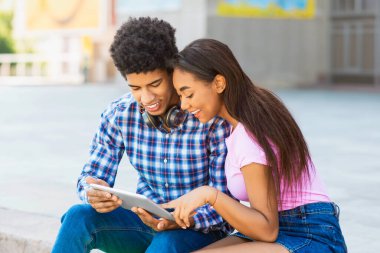 Afrika kökenli Amerikalı genç bir erkek ve kız güneşli bir günde dışarıdaki merdivenlerde oturuyorlar. Kız pembe gömlek ve kot şort giyerken adam mavi ekose gömlek ve kot pantolon giyiyor..