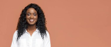 Uzun, siyah kıvırcık saçlı ve beyaz yakalı bir gömlek giyen Afro-Amerikalı genç bir kadın şeftali rengi bir arka plana gülümsüyor..