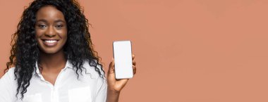 Uzun kıvırcık siyah saçlı, Afro-Amerikalı bir kadın beyaz yakalı bir gömlek giyiyor ve şeftali renkli bir arka plan önünde akıllı telefonu tutarken geniş bir gülümseme sergiliyor..