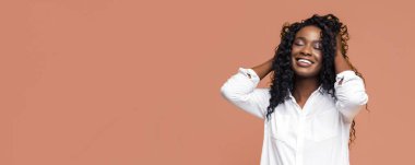Uzun kıvırcık saçlı siyah bir kadın kameraya gülümsüyor. O, beyaz düğmeli bir gömlek giyiyor ve elleri başının arkasında. Arka plan bronz bir renk..