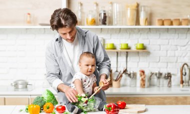 Bir baba ve bebeği mutfakta sebze hazırlıyorlar. Baba, tahta kaşıkla bir kase yeşili karıştırırken bebeği tutuyor. Bebek sebzelere ilgiyle bakıyor.