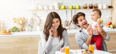 Genç bir kadın mutfak masasında oturuyor, telefonda konuşuyor ve kocası bebeğini havada tutarken not alıyor. Bebek babasına bakıyor ve gülümsüyor.